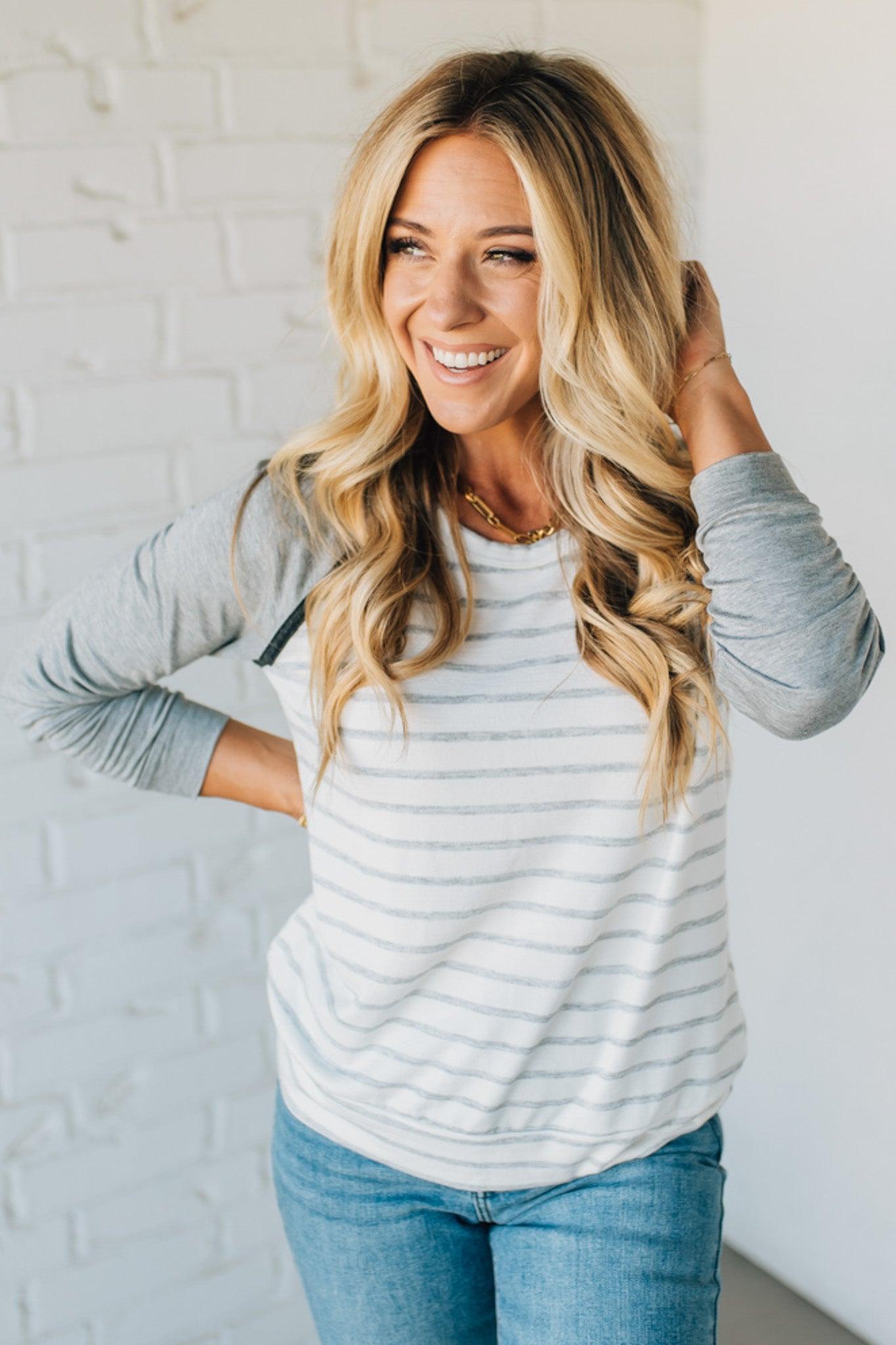 Woman wearing a gray and white striped sweater with blue jeans against a white brick wall.