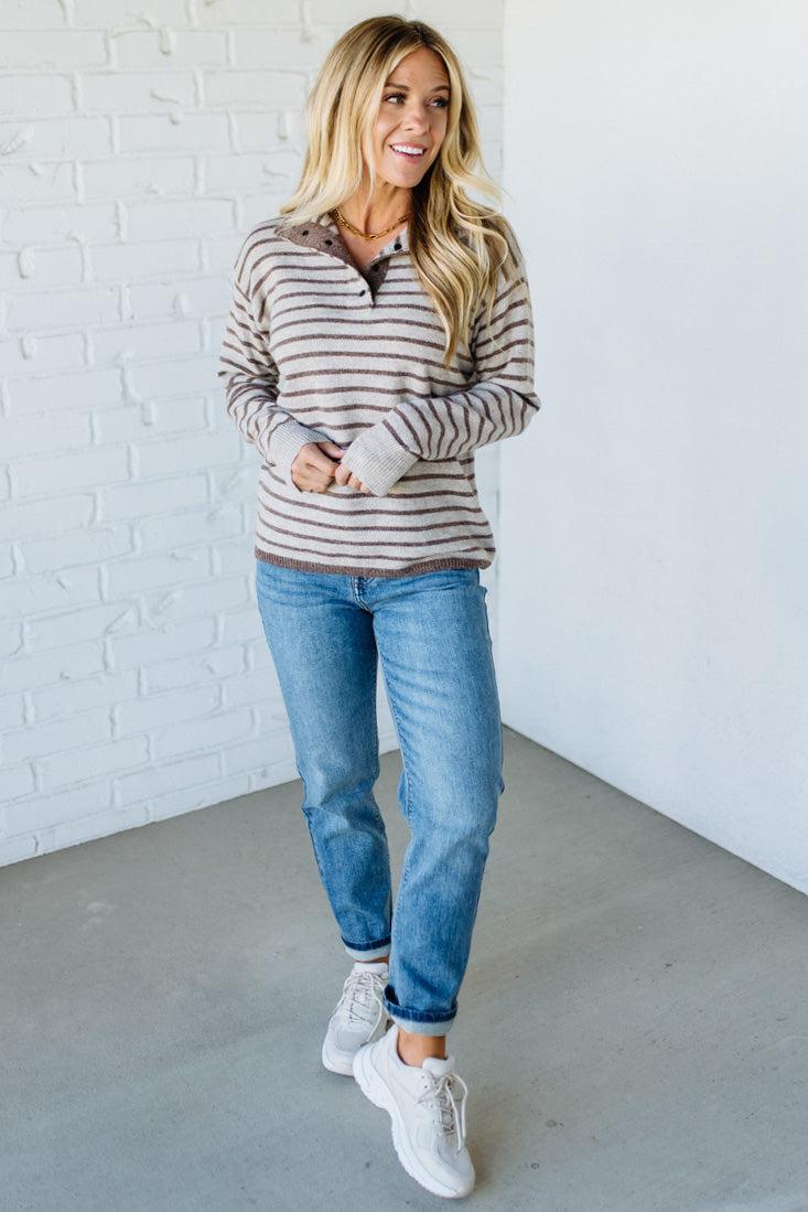 Woman wearing a striped sweater, jeans, and white sneakers against a white brick wall.