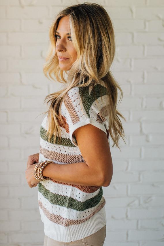 Blonde woman wearing a cream, olive green, mocha and rust striped sweater with jacquard loose weave pattern.