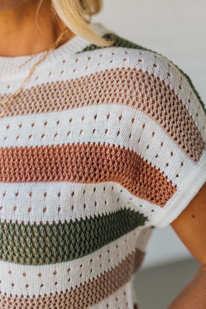 Blonde woman wearing a cream, olive green, mocha and rust striped sweater with jacquard loose weave pattern.
