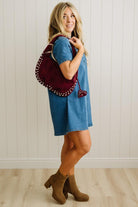 Woman in a blue dress holding a red backpack against a white paneled wall.