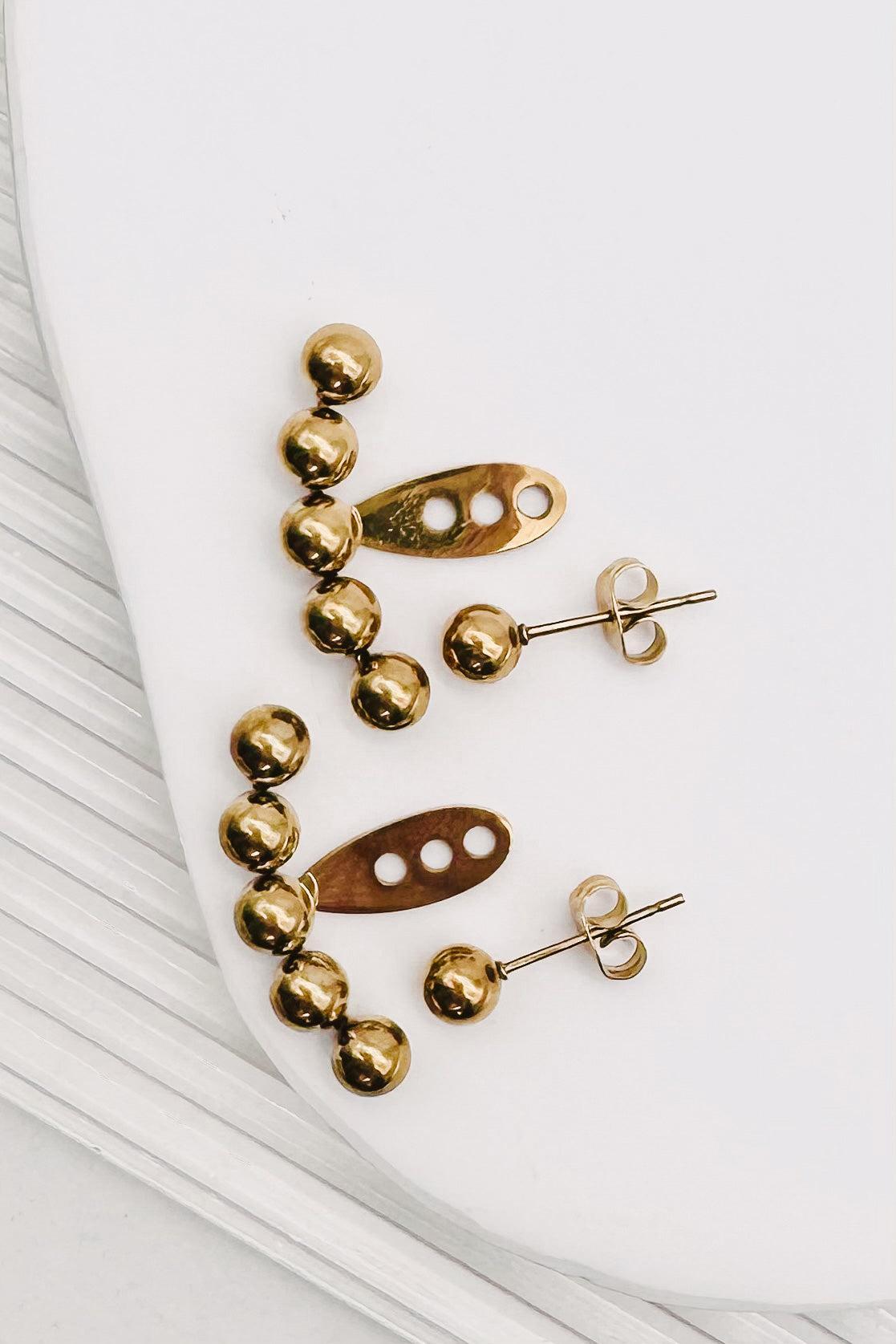 A pair of gold-toned statement earrings with a curved row of polished beads and a central abstract cutout bar, displayed on a white surface.