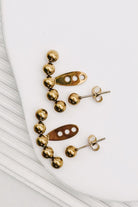 A pair of gold-toned statement earrings with a curved row of polished beads and a central abstract cutout bar, displayed on a white surface.