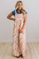 Woman wearing a floral overalls with a blue top against a white wall.