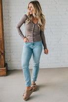 Woman wearing a brown cardigan, blue jeans, and sandals against a white brick wall.