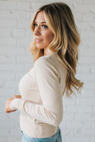 Woman wearing a beige sweater and blue jeans against a white brick wall.