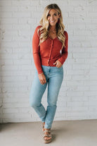 Woman wearing a red sweater and light blue jeans standing against a white brick wall.