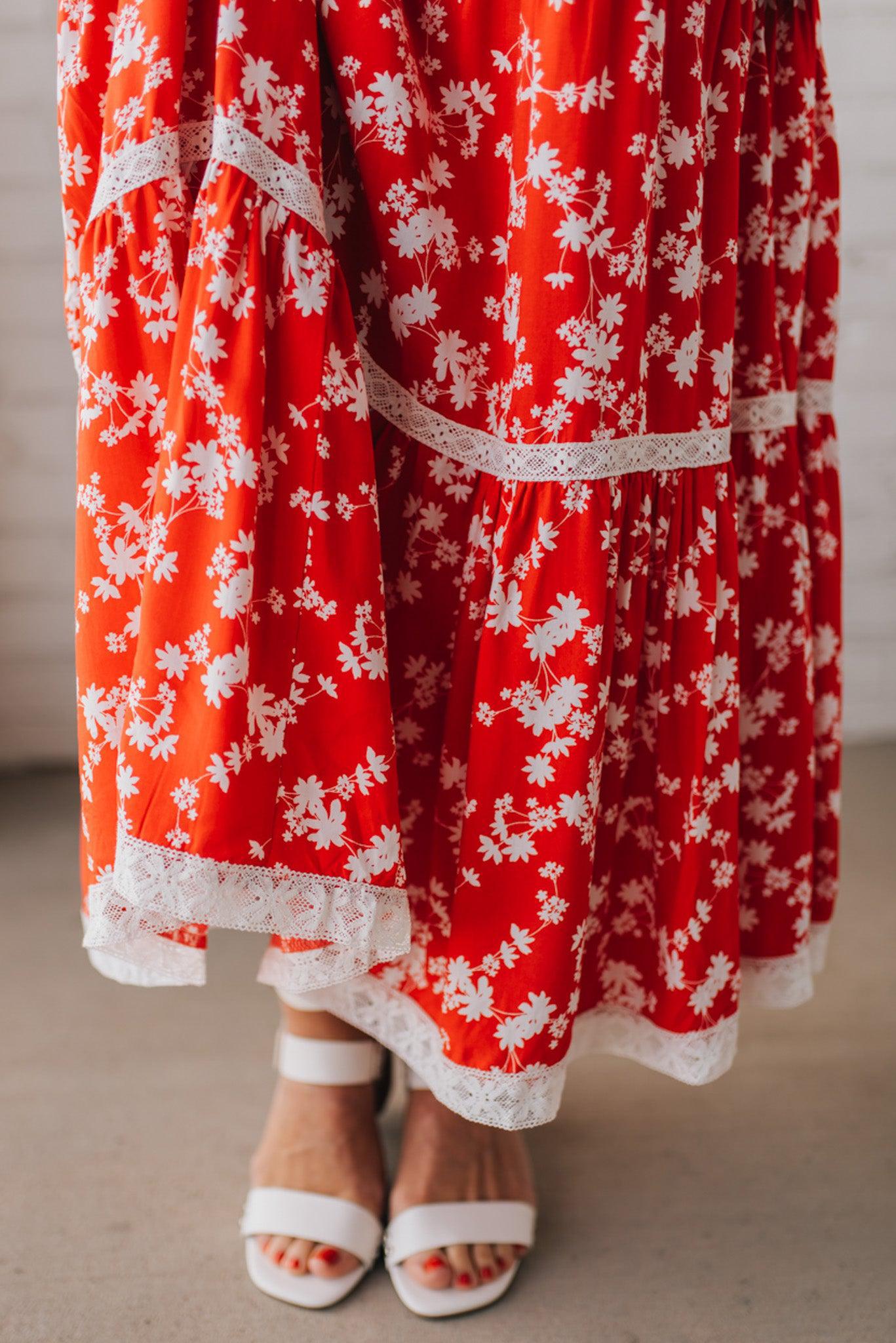 BLONDE WOMAN WEARING A RED FLORAL PRINT SLEEVELESS MAXI FEATURES: TIERED SKIRT PANEL WITH LACE INSET DETAIL, SMOCKED BACK, HIDDEN SIDE ZIPPER, THICK STRAPS WITH SCALLOPED LACE EDGES, SIDE POCKETS AND FULL LINING.