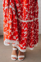 BLONDE WOMAN WEARING A RED FLORAL PRINT SLEEVELESS MAXI FEATURES: TIERED SKIRT PANEL WITH LACE INSET DETAIL, SMOCKED BACK, HIDDEN SIDE ZIPPER, THICK STRAPS WITH SCALLOPED LACE EDGES, SIDE POCKETS AND FULL LINING.