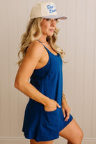 Woman wearing a blue romper and beige cap with text against a neutral background
