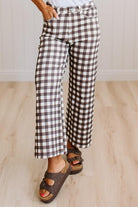 A person wearing brown and white gingham cropped flare pants with brown platform sandals, standing on a light wood floor against a white paneled wall.