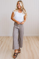 A woman modeling brown and white gingham wide-leg cropped pants, paired with a white t-shirt and brown slide sandals, standing in a room with light wood floors and white paneled walls.