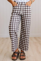 A person wearing brown and white gingham cropped wide-leg pants, paired with a white top and brown platform sandals, standing on a light wood floor against a white paneled wall.