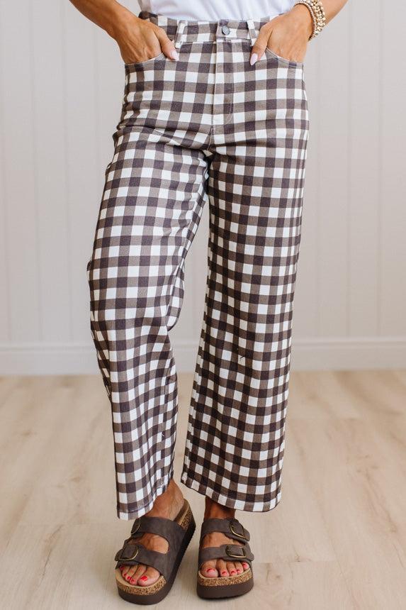A person wearing brown and white gingham cropped wide-leg pants, paired with a white top and brown platform sandals, standing on a light wood floor against a white paneled wall.