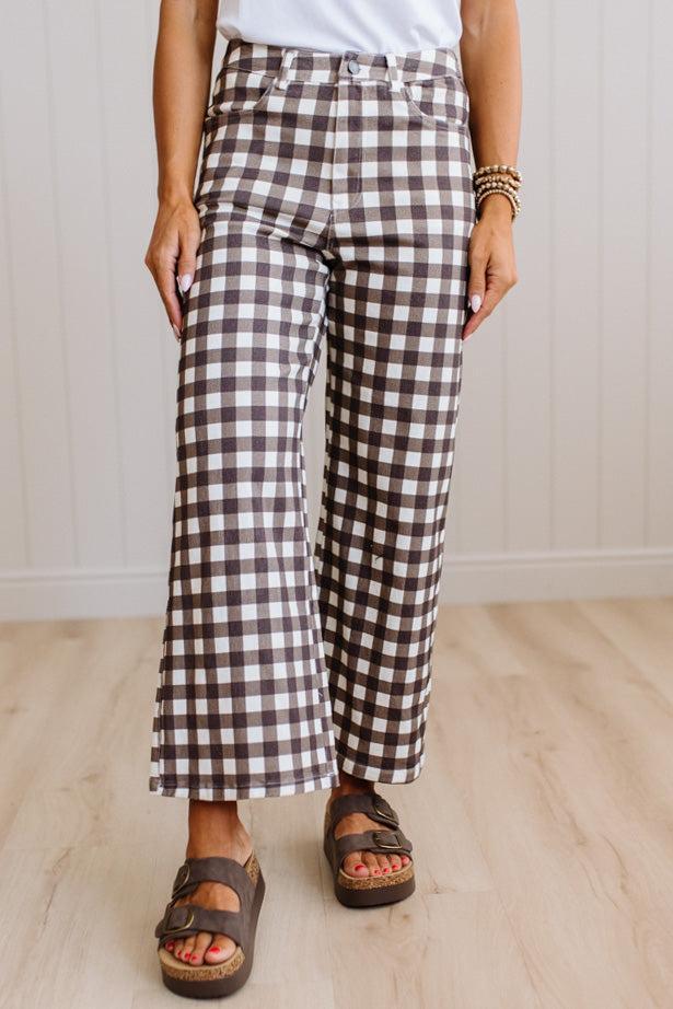 A person wearing brown and white gingham wide-leg cropped pants with a white top and brown platform sandals, standing on a light wood floor against a white paneled wall.