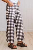A person wearing brown and white gingham wide-leg cropped pants, paired with brown slide sandals and a white top, standing in a room with light wood floors and white paneled walls.