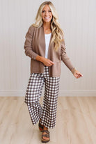 A woman modeling a brown cardigan over a white top and brown-and-white gingham wide-leg pants, standing in a bright room with light wood floors and white paneled walls.