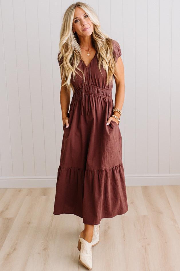 Blonde woman modeling a brown v-neck mid-length dress with tie front tassels and side pockets. 