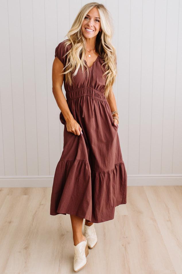 Blonde woman modeling a brown v-neck mid-length dress with tie front tassels and side pockets. 