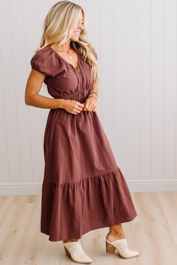 Blonde woman modeling a brown v-neck mid-length dress with tie front tassels and side pockets. 