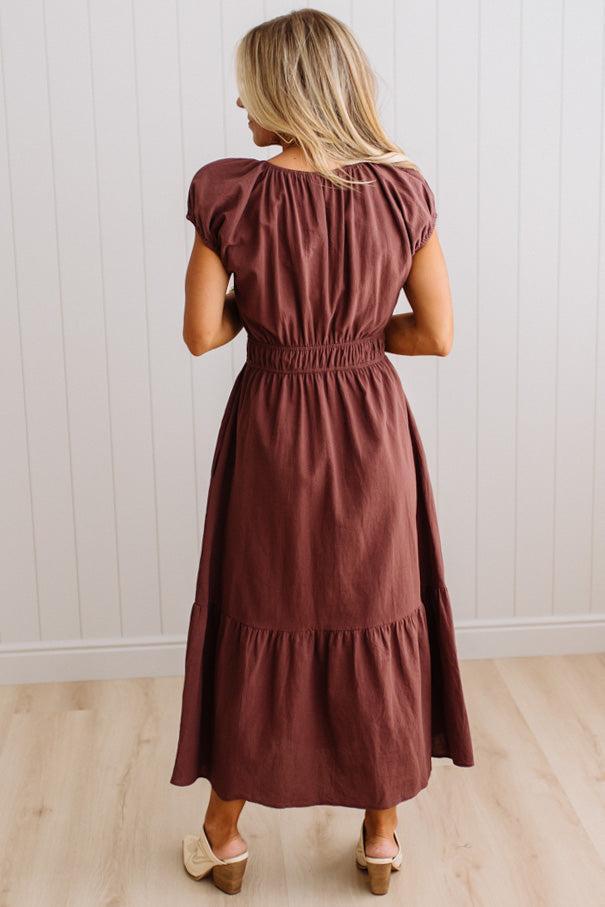 Blonde woman modeling a brown v-neck mid-length dress with tie front tassels and side pockets. 
