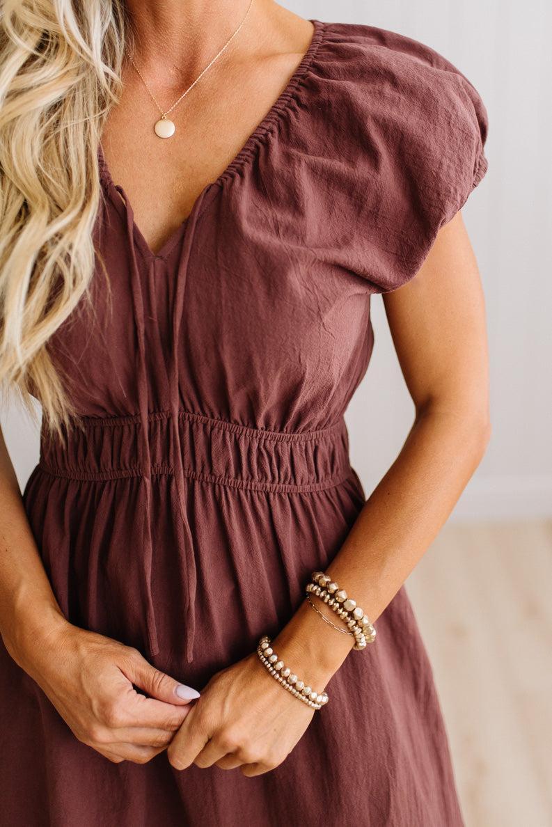 Blonde woman modeling a brown v-neck mid-length dress with tie front tassels and side pockets. 