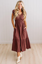 Blonde woman modeling a brown v-neck mid-length dress with tie front tassels and side pockets. 