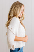 BLONDE WOMAN WEARING DARK JEANS IN A BRIGHTLY LIT STUDIO WITH A SOLID SWEATER FEATURING SCALLOP TRIM AND BUTTONS UP THE SLEEVE IN THE COLOR HEATHERED OATMEAL.