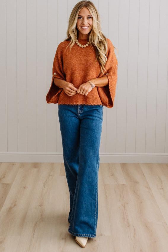 BLONDE WOMAN WEARING DARK JEANS IN A BRIGHTLY LIT STUDIO WITH A SOLID SWEATER FEATURING SCALLOP TRIM AND BUTTONS UP THE SLEEVE IN THE COLOR HEATHERED RUST.