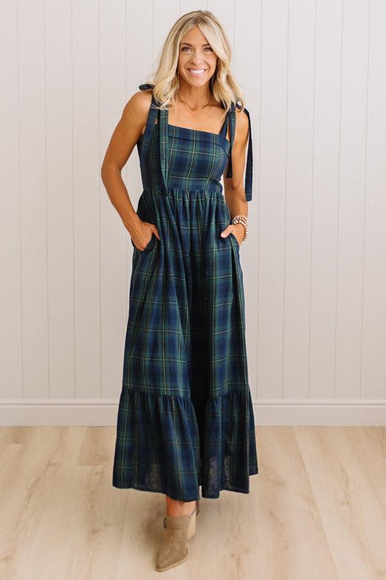 A woman modeling a dark green and navy plaid maxi dress with tie-shoulder straps and side pockets, standing against a white paneled wall and light wood floor.