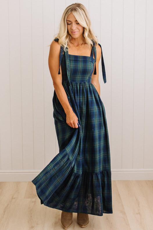 A woman modeling a long plaid maxi dress with tie straps and side pockets, standing in front of a white paneled wall and light wood floor.