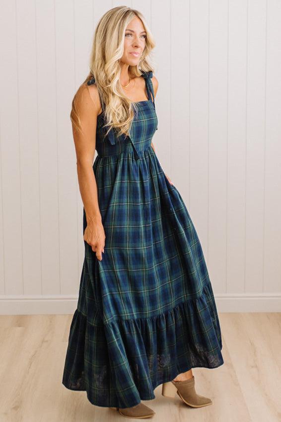 A woman modeling a sleeveless, tiered maxi dress in a dark blue and green plaid pattern, paired with tan heeled boots, standing against a white paneled wall and light wood floor.