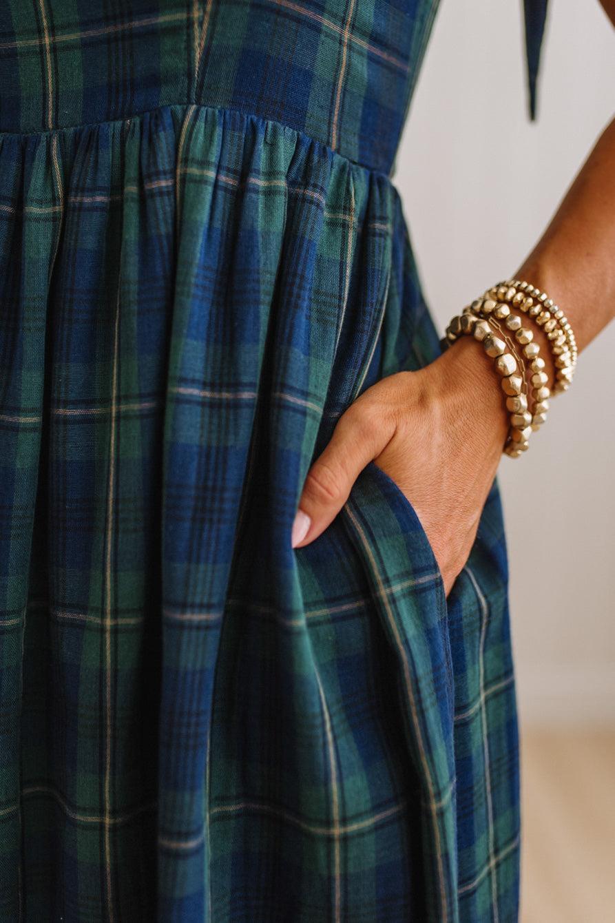 Close-up of a blue and green plaid dress with a side pocket, shown on a model whose hand is tucked inside the pocket. The model is wearing stacked gold beaded bracelets.
