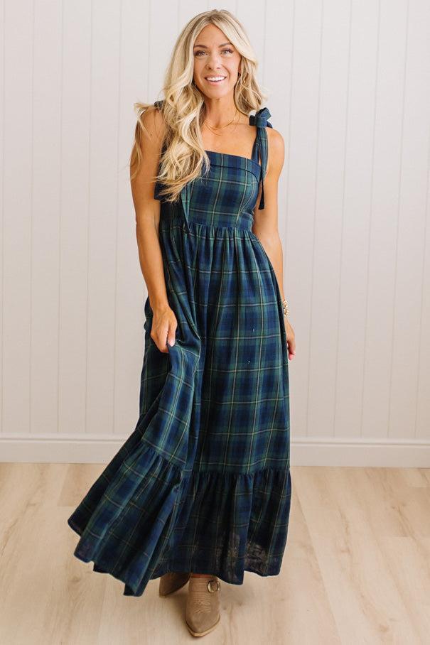 A woman modeling a sleeveless navy blue plaid maxi dress with tie straps and a tiered skirt, standing in front of a white paneled wall and light wood floor.