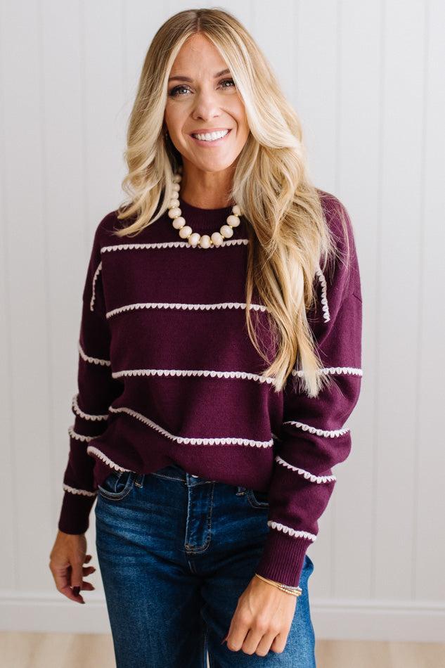 BLONDE WOMAN WEARING DARK JEANS IN A BRIGHTLY LIT STUDIO MODELING A CREW NECK, LONG SLEEVE, BLANKET STITCH STRIPED SWEATER IN DARK PLUM.