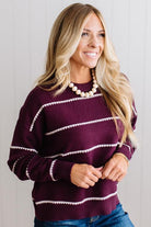 BLONDE WOMAN WEARING DARK JEANS IN A BRIGHTLY LIT STUDIO MODELING A CREW NECK, LONG SLEEVE, BLANKET STITCH STRIPED SWEATER IN DARK PLUM.