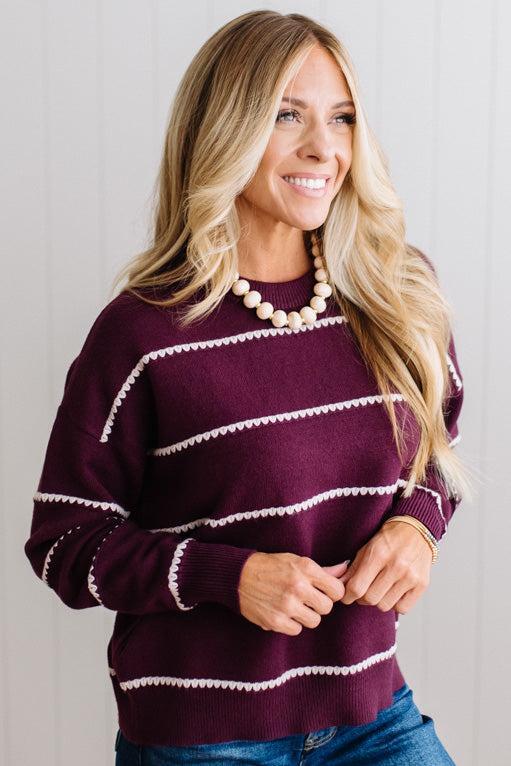 BLONDE WOMAN WEARING DARK JEANS IN A BRIGHTLY LIT STUDIO MODELING A CREW NECK, LONG SLEEVE, BLANKET STITCH STRIPED SWEATER IN DARK PLUM.