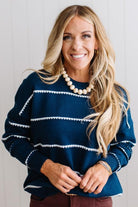 BLONDE WOMAN WEARING BROWN JEANS IN A BRIGHTLY LIT STUDIO MODELING A CREW NECK, LONG SLEEVE, BLANKET STITCH STRIPED SWEATER IN DARK TEAL.