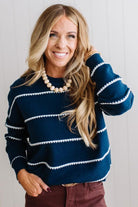 BLONDE WOMAN WEARING BROWN JEANS IN A BRIGHTLY LIT STUDIO MODELING A CREW NECK, LONG SLEEVE, BLANKET STITCH STRIPED SWEATER IN DARK TEAL.