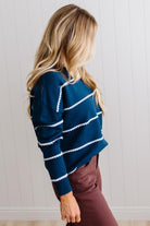 BLONDE WOMAN WEARING BROWN JEANS IN A BRIGHTLY LIT STUDIO MODELING A CREW NECK, LONG SLEEVE, BLANKET STITCH STRIPED SWEATER IN DARK TEAL.