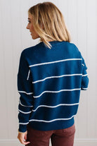 BLONDE WOMAN WEARING BROWN JEANS IN A BRIGHTLY LIT STUDIO MODELING A CREW NECK, LONG SLEEVE, BLANKET STITCH STRIPED SWEATER IN DARK TEAL.
