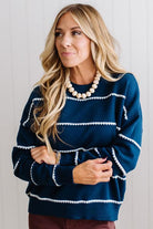 BLONDE WOMAN WEARING BROWN JEANS IN A BRIGHTLY LIT STUDIO MODELING A CREW NECK, LONG SLEEVE, BLANKET STITCH STRIPED SWEATER IN DARK TEAL.