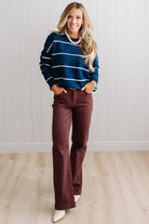 BLONDE WOMAN WEARING BROWN JEANS IN A BRIGHTLY LIT STUDIO MODELING A CREW NECK, LONG SLEEVE, BLANKET STITCH STRIPED SWEATER IN DARK TEAL.