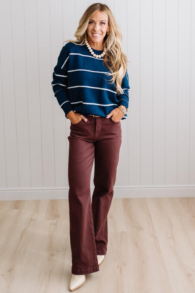 BLONDE WOMAN WEARING BROWN JEANS IN A BRIGHTLY LIT STUDIO MODELING A CREW NECK, LONG SLEEVE, BLANKET STITCH STRIPED SWEATER IN DARK TEAL.