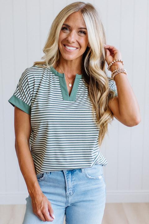 Woman wearing a green striped shirt and light jeans standing in a room with light wood flooring and white walls.