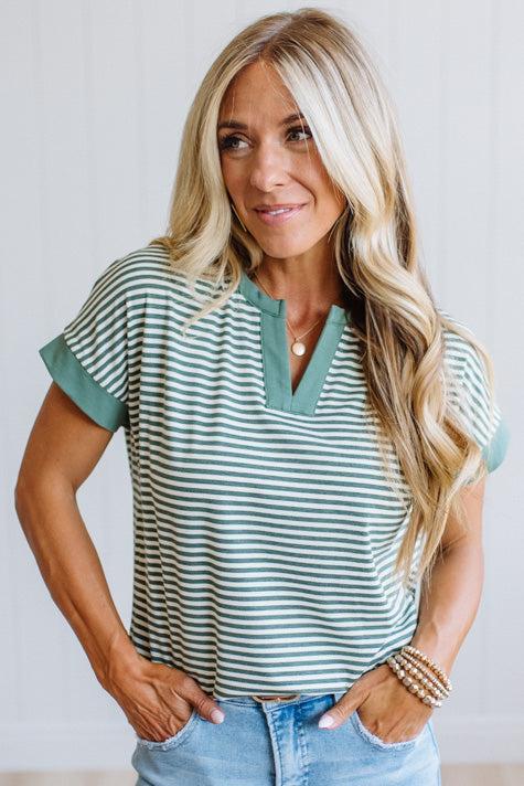 Woman wearing a green striped shirt and light jeans standing in a room with light wood flooring and white walls.