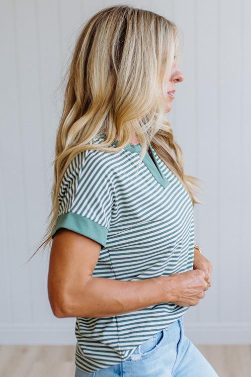 Woman wearing a green striped shirt and light jeans standing in a room with light wood flooring and white walls.
