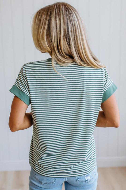 Woman wearing a green striped shirt and light jeans standing in a room with light wood flooring and white walls.