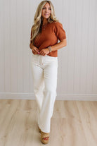 Woman wearing a brown ribbed sweater and white pants standing against a light wooden paneled wall.