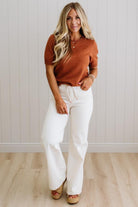 Woman wearing a rust-colored top and white pants standing against a light wooden paneled wall.